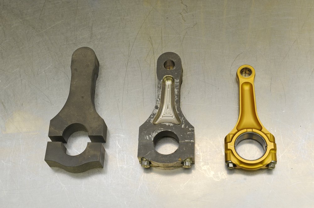 L&rsquo;immagine mostra una biella in lega di titanio in tre fasi del processo di fabbricazione. Il pezzo grezzo di partenza (a sinistra) &egrave; ottenuto dalla lastra, mediante taglio ad acqua
