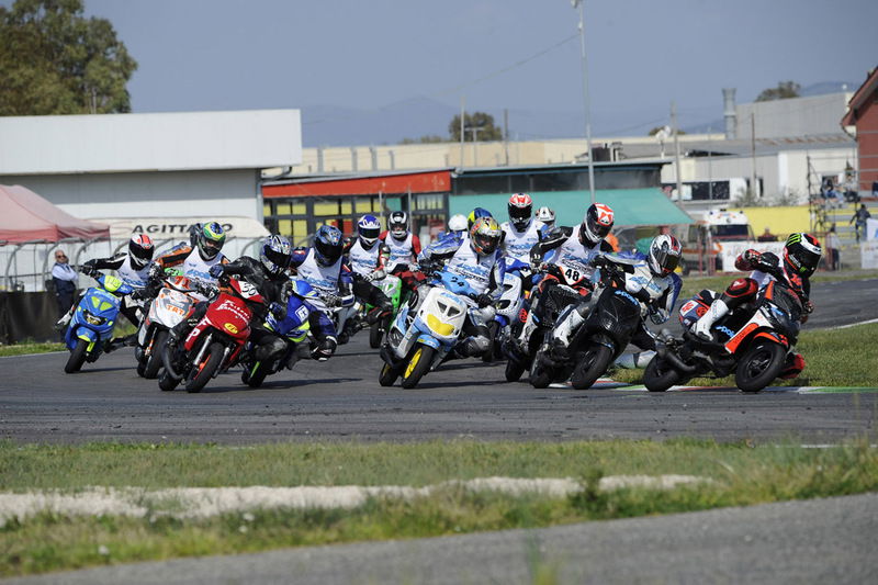 Grande partecipazione di piloti nella Scooter 70 Evolution Amatori con due Gare tiratissime e ricche di spettacolo. Qui il gruppone affronta la prima curva della rinnovata pista di Latina con il vincitore assoluto Guido Conforti (48) in terza posizione, mentre al comando &egrave; Rocco Russo (14) che finir&agrave; sul secondo gradino del podio davanti a Davide Inversini (24), terzo di giornata.